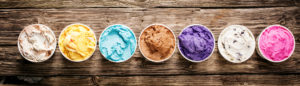 Row of assorted flavors and colors of gourmet Italian ice cream served in plastic takeaway tubs on a rustic wooden table, horizontal banner format with copyspace