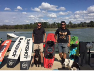 Two young men in t-shirts and shorts stand next to four large wakeboards. A large lake is behind them, with trees in the far background. A cute dog sits at the feet of one man, looking at the camera.