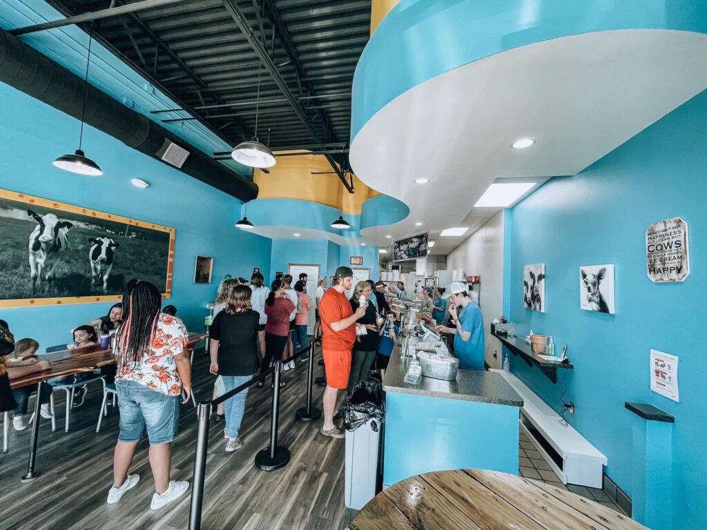 Wide interior view of a bright ice cream shop with turquoise walls, customers standing in line, employees behind a service counter, and framed cow artwork on the walls.