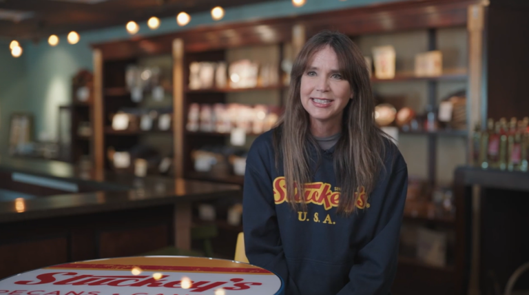 Person wearing a Stuckey’s U.S.A. sweatshirt seated indoors at a table, with shelves of packaged food products and warm string lighting in the background.