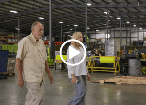Older man and woman walk side by side through a large warehouse filled with industrial equipment, pallets, and workstations.