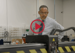 Man wearing glasses and a dress shirt with a patterned tie examines industrial equipment on a manufacturing floor, focused on the machinery in front of him.