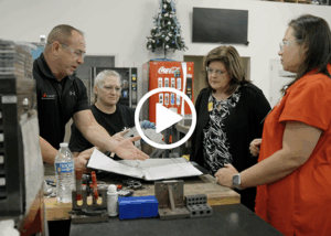 Four adults wearing safety glasses stand around a workbench in an industrial workshop as a man gestures to documents and metal parts, discussing a manufacturing process.