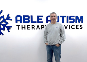 Idris Demirci stands in front of a white wall bearing the name and logo of his business, Able Autism Therapy Services.