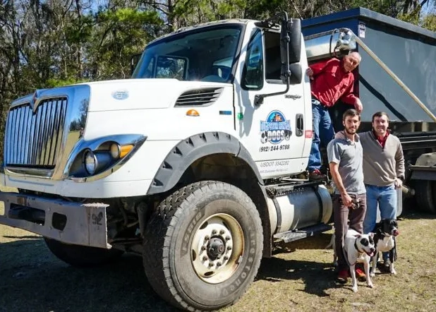 Three people and two dogs are standing in front of the large white truck with a portable dumpster on the back. One person is standing on the truck's step, while the other two people, Max Lemaigre and Sean Dane-Kellogg, and are standing on the ground next to each other, holding leashes attached to the dogs.