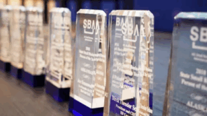 Glass awards with blue bases, engraved with the SBA logo and inscriptions, lined up on a table with a blurred blue background.