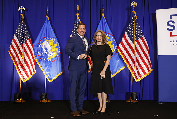 Patrick Wells, owner and president of Piedmont Landscape Management, stands on stage in front of blue curtain to receive SBA award plaque from woman in black dress.