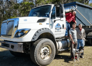 Three people and two dogs are standing in front of the large white truck with a portable dumpster on the back. One person is standing on the truck's step, while the other two people, Max Lemaigre and Sean Dane-Kellogg, and are standing on the ground next to each other, holding leashes attached to the dogs.