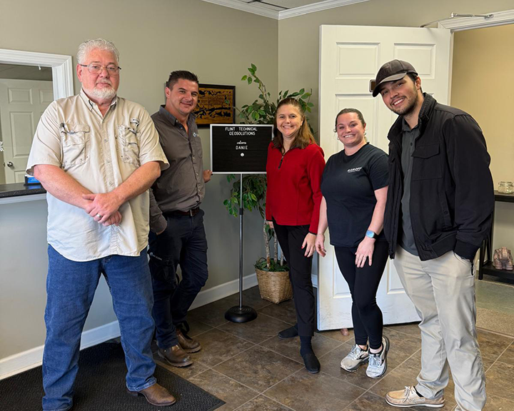 A group of five people standing in an office-like environment, positioned near a sign that reads ‘Flint Technical Geosolutions.’ The individuals are dressed casually, and there is a potted plant and some office furniture visible in the background.