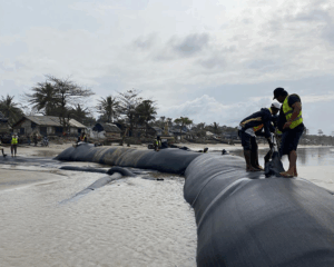 Workers in high-visibility vests install large black textile barriers filled with stone and sand on a beach, with palm trees and buildings in the background under a cloudy sky.