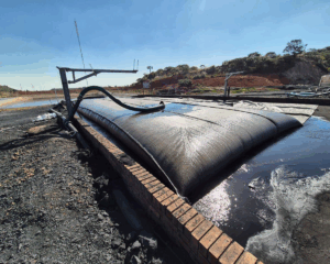 : A large, inflated black bag-like structure rests on a brick-lined platform. Hoses are connected to the structure, and the surrounding area is wet. In the background, there are hills with vegetation and some industrial equipment under a clear blue sky.