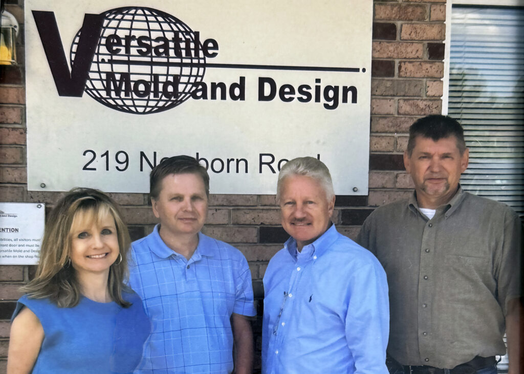 Four people stand, a woman and three men, in front of a brick wall with a sign that reads 'Versatile Mold and Design' and '219 Newborn Road.’