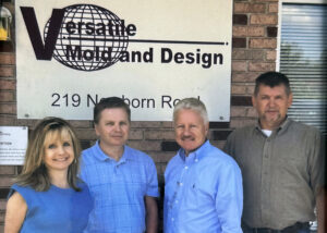 Four people stand, a woman and three men, in front of a brick wall with a sign that reads 'Versatile Mold and Design' and '219 Newborn Road.’