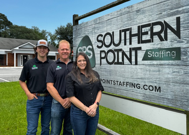 Three people in black company shirts stand in front of a 'Southern Point Staffing' sign on a grassy area with building in the background.