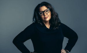 A woman with long, dark hair is standing with hands on hips, wearing a black sweater. The background is a plain gray color.