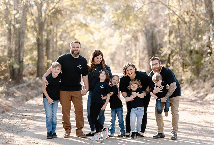 Group of nine people, including adults and children, wearing matching black shirts with a white logo, standing on a sunlit dirt path in a wooded area surrounded by tall trees.