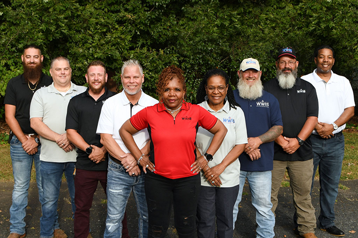 Nine people posing outdoors in front of greenery, wearing casual 'WWIS' logo shirts. The person in the center front wears a red polo shirt, while others wear black, white, or blue shirts.