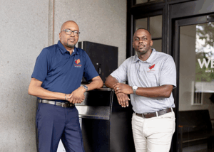 Two men in branded polo shirts lean against a valet stand outside a building with a glass door showing partial signage.