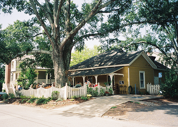 A yellow house with a white picket fence and a large tree in front. People are sitting on the porch and in the yard. A driveway leads to the house, with another building visible in the background. The scene is sunny with lush green trees providing shade.