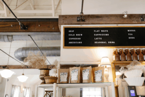 Coffee shop interior with a hanging menu board listing drinks including drip coffee, cold brew, espresso, and tea. Below the board are shelves displaying coffee bean bags and decorative items.
