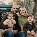 Family of four sitting outdoors in front of a large tree. Father holds a baby, mother sits with her arm around their young child. Cozy setup with outdoor furniture and trees in the background.