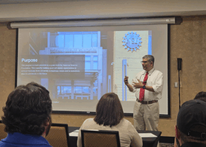 A man in a white shirt and red tie presents to an audience seated at tables. Behind him, a slide displays the National Science Foundation (NSF) logo and building, with text explaining a grant.
