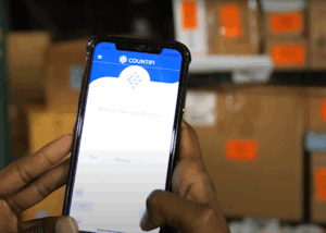 : A person holds a smartphone displaying the COUNTIFI app in use. In the background, several cardboard boxes with orange labels are visible, indicating a warehouse or storage setting.