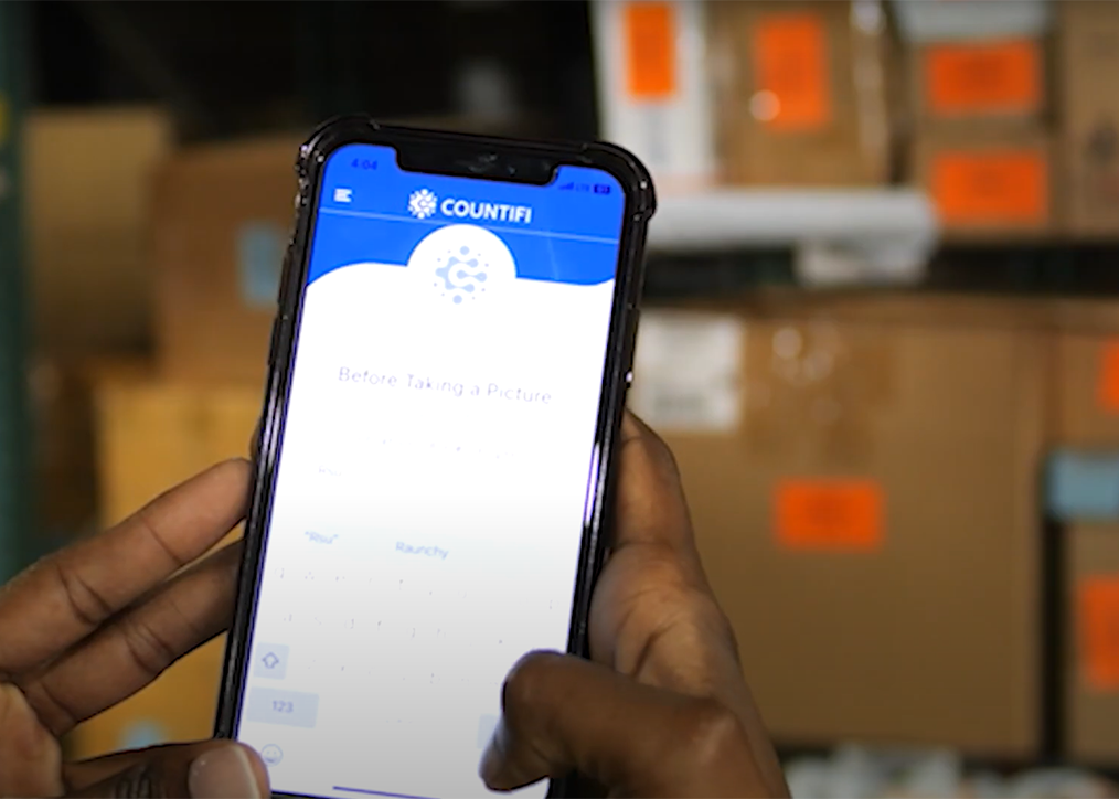 : A person holds a smartphone displaying the COUNTIFI app in use. In the background, several cardboard boxes with orange labels are visible, indicating a warehouse or storage setting.