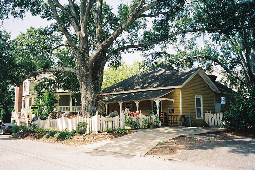 A yellow house with a white picket fence and a large tree in front. People are sitting on the porch and in the yard. A driveway leads to the house, with another building visible in the background. The scene is sunny with lush green trees providing shade.