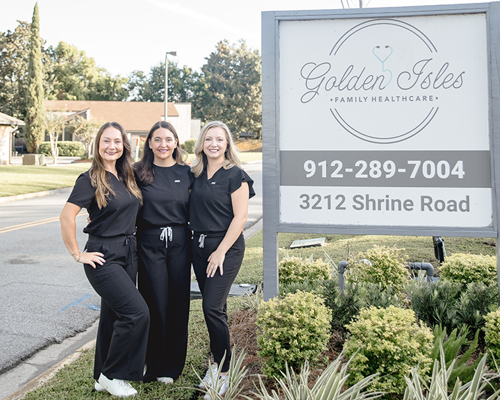 Three individuals dressed in black medical scrubs stand smiling next to a large sign for Golden Isles Family Healthcare. The background shows a road, greenery, and buildings.