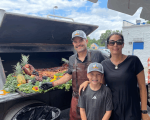 Three people stand in front of a barbecue grill filled with grilled meats, fruits, and vegetables.