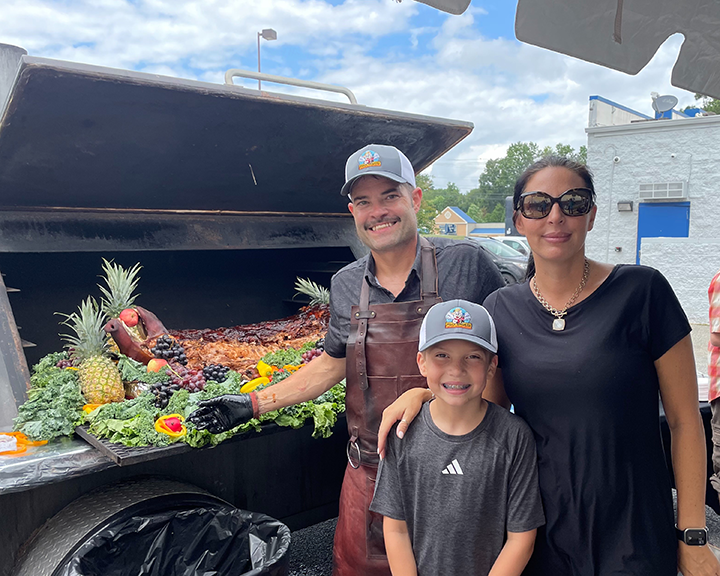 Three people stand in front of a barbecue grill filled with grilled meats, fruits, and vegetables.