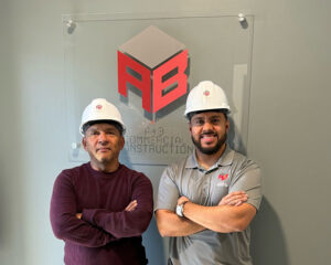 Two construction professionals wearing white safety helmets stand confidently in front of a gray wall with the "AB Commercial Construction" logo. One wears a maroon long-sleeve shirt, the other a gray polo with the company’s logo. Both are smiling with arms crossed.