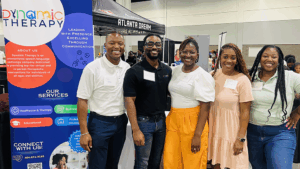 Group of five people standing together at an indoor event next to a tall banner for “Dynamic Therapy.” The banner includes text about services such as healthcare and therapy, business, and educational support. Behind them are other booths.