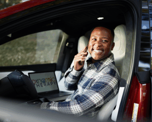 Person sitting in the driver’s seat of a car, holding a smartphone to their ear. A laptop with the words “Dynamic Therapy” on the screen is placed on the passenger seat. The car door is open, and the interior appears modern with light-colored seats.
