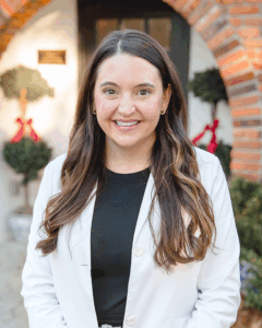 Smiling woman in a white coat stands outside a brick building with festive potted plants.