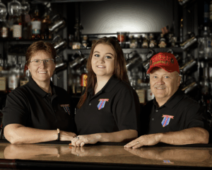 Three individuals wearing matching black collared shirts with a colorful embroidered logo stand behind a polished bar counter. The background features shelves filled with bottles, glassware, and metallic mugs, creating a classic bar setting. One person on the right is wearing a bright red cap with yellow lettering.