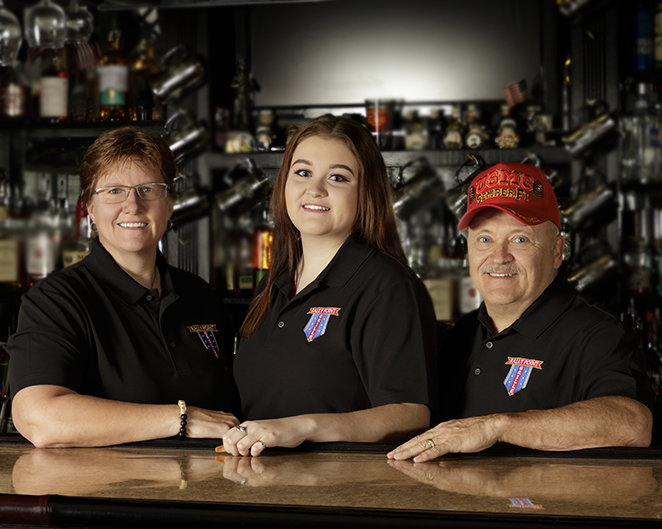 Three individuals wearing matching black collared shirts with a colorful embroidered logo stand behind a polished bar counter. The background features shelves filled with bottles, glassware, and metallic mugs, creating a classic bar setting. One person on the right is wearing a bright red cap with yellow lettering.