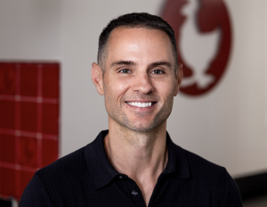 Person wearing a dark collared shirt, positioned indoors against a background featuring a red tiled section and a large circular red logo with a white rooster silhouette.