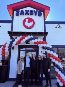 Five people standing in front of a Zaxbys restaurant entrance decorated with an arch of red, white, and black balloons. The building features a white exterior with dark trim, a red roof accent, and a large Zaxbys logo with a chicken silhouette above the door.