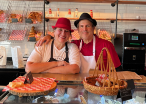 Two people wearing aprons and hats stand behind a bakery counter with baskets of bagels and pastries lined with red-and-white checkered paper. Behind them, shelves display bagels and bread, along with decorative items and nameplates.