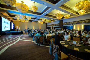 A large conference room filled with attendees seated at round tables, facing a stage with a speaker presenting. The stage has a large screen displaying the text “Importance of Reduce before Recycle” alongside graphics. The room features high ceilings with multiple ornate chandeliers, warm lighting, and patterned carpet.