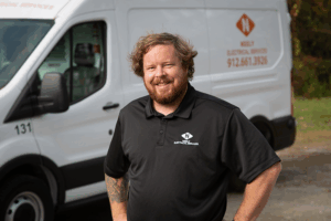 Man wearing a black polo shirt with the Neely Electrical Services logo standing in front of a white service van. The van displays the company name, logo, and phone number on its side.