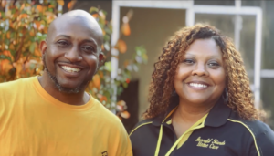 Two people stand side by side outdoors in front of autumn-colored foliage. One person wears a bright yellow T‑shirt, and the other wears a black collared shirt with yellow trim that reads “Graceful Hands Home Care.”