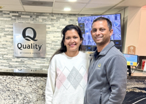 Two people stand together in the lobby of a hotel, positioned in front of a stone-textured wall with a large “Quality by Choice Hotels” sign. Behind They are standing near a granite-topped front desk, and one person is wearing a light-colored sweater while the other is wearing a zip-up jacket with the hotel logo.