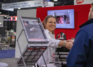 Representative at a trade show booth shaking hands with an attendee next to food processing machinery and a product display screen.
