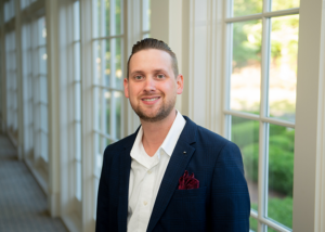 Person wearing a navy plaid blazer and white shirt stands indoors along a hallway of large windows with greenery visible outside.