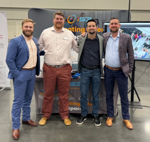 Four people stand side by side in front of an iS3 Tech exhibition booth at an indoor event, with a branded backdrop and display screen behind them.