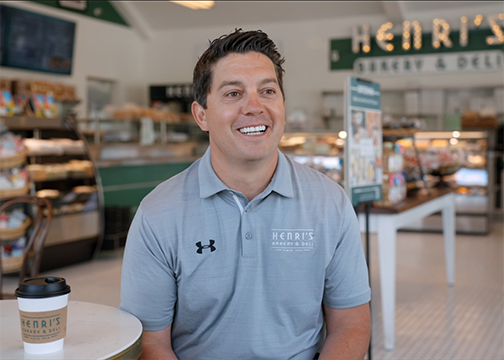Person in a gray Henri's Bakery & Deli polo shirt sits at a table with a coffee cup, surrounded by bakery displays and shelves. A large "Henri's" sign is visible in the background.