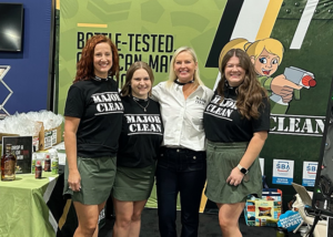 Four people stand side by side at a trade show booth, posing in front of a green backdrop that reads “Battle-Tested Performance.” Three individuals wear black “MAJOR CLEAN” T-shirts with green skirts, while the person in the center wears a white button-down shirt. A table with branded products and signage is visible to the left, and promotional materials and boxes appear on the right, reinforcing the product-focused display.